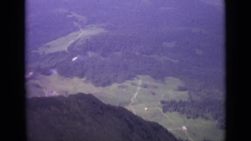 Aerial Swiss valley view from 1962 showing meadow patches, forest, pale tracks, and a dark hillside foreground.