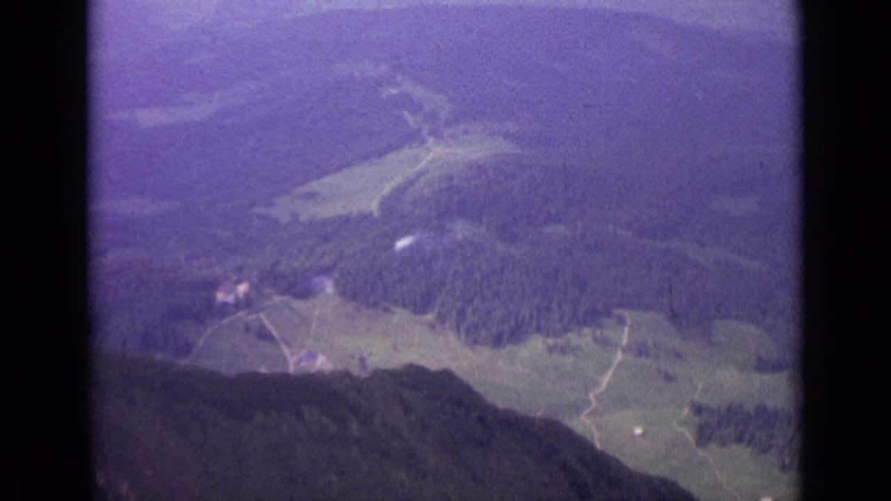 1962 Swiss aerial home-movie frame showing fields, woodland, tracks, and small distant structures in a hazy valley.