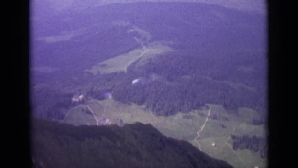 1962 Swiss aerial home-movie frame showing fields, woodland, tracks, and small distant structures in a hazy valley.