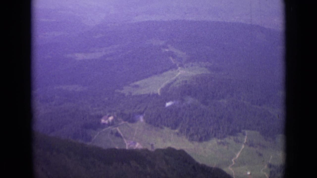 Soft 8mm aerial view over Switzerland in 1962, with forest, meadow clearings, pale paths, and distant hillside terrain.