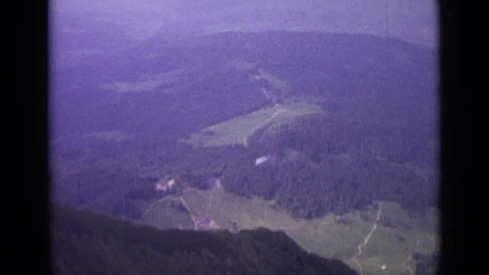 Soft 8mm aerial view over Switzerland in 1962, with forest, meadow clearings, pale paths, and distant hillside terrain.