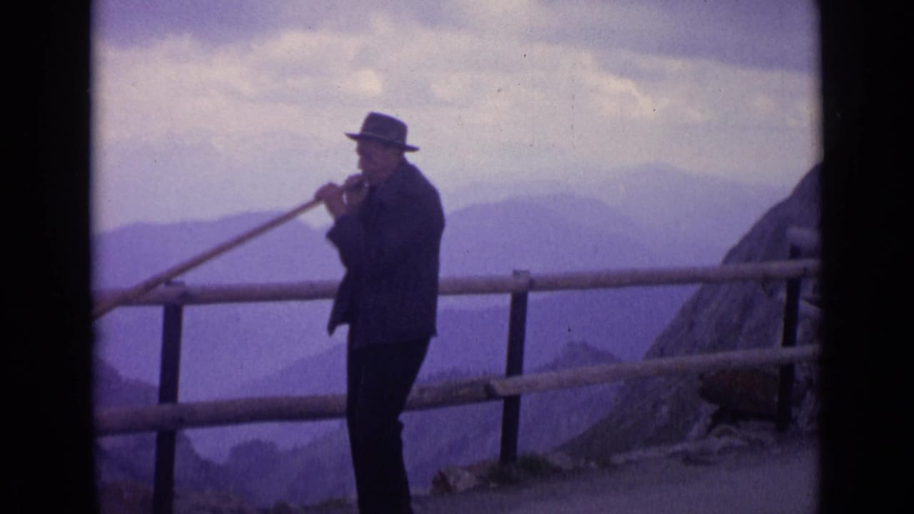 1962 Switzerland 8mm frame of a suited man in a brimmed hat playing a long wooden horn at an alpine overlook.