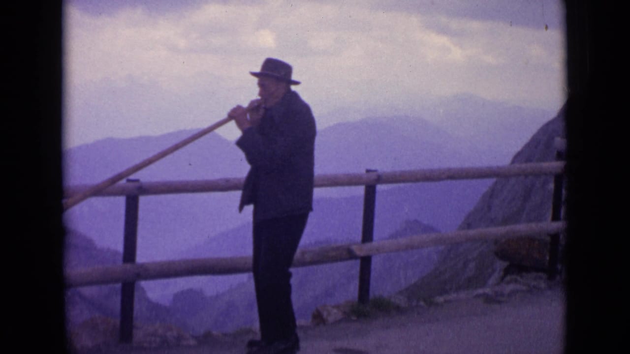 1962 Switzerland 8mm frame showing a man performing with a long wooden horn at a mountain viewpoint.