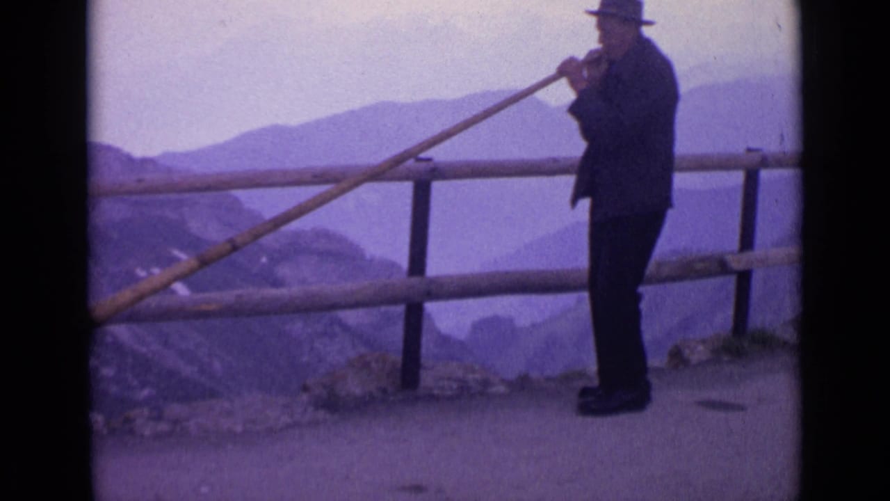 1962 Switzerland 8mm frame of the horn player in profile near a wooden rail, with hazy alpine ridges beyond.