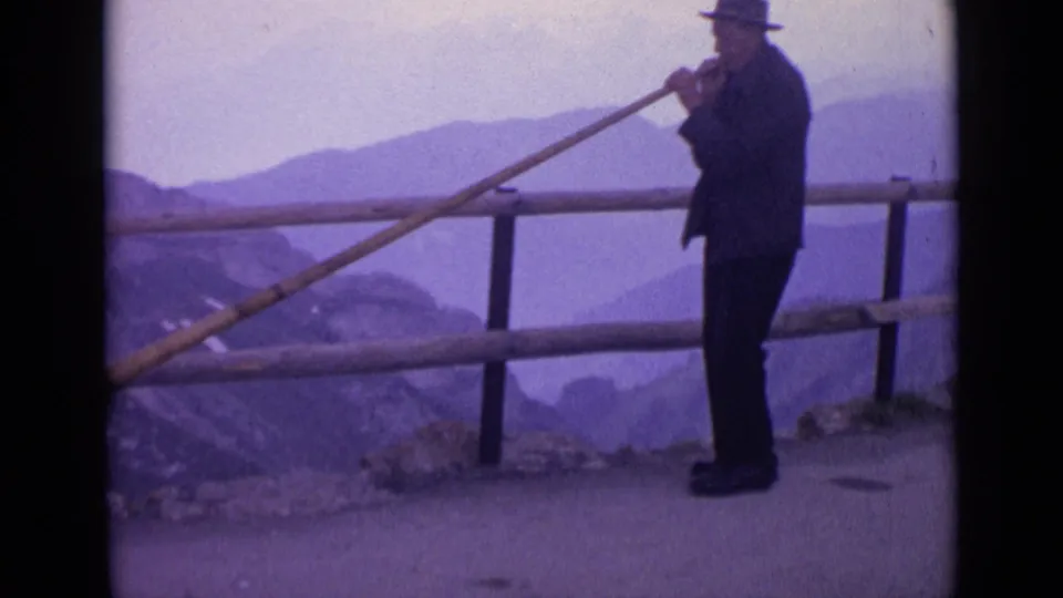 1962 Switzerland 8mm frame of the horn player in profile near a wooden rail, with hazy alpine ridges beyond.
