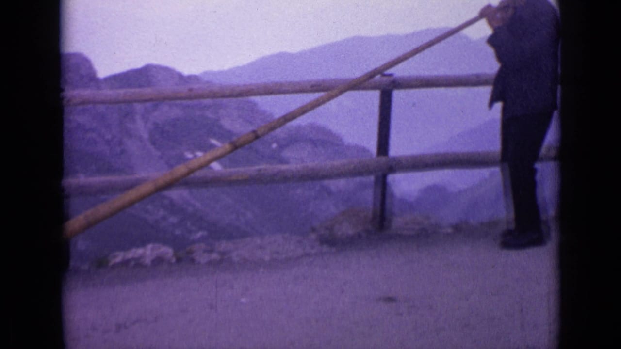 1962 Switzerland home-movie frame showing a man at an alpine overlook holding a long wooden horn beside a railing and hazy peaks.