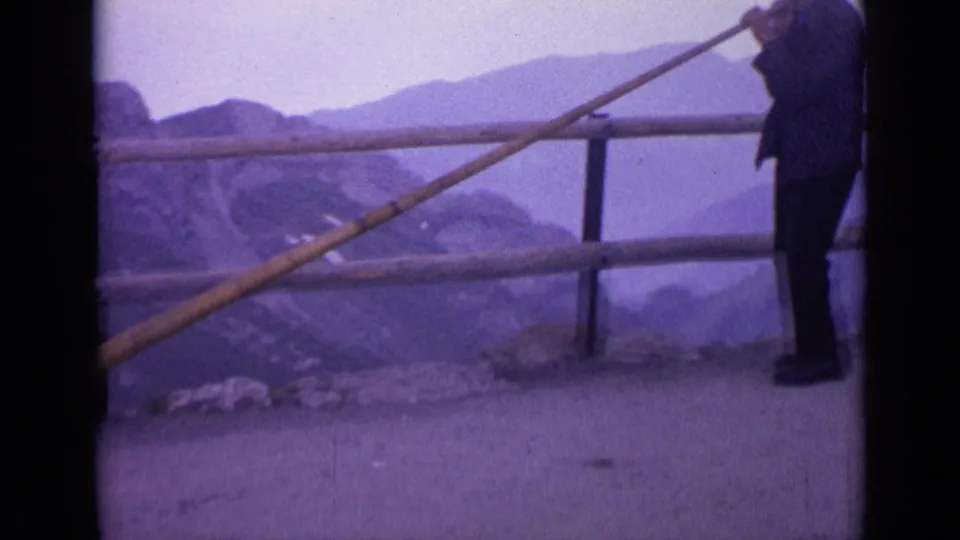 1962 Switzerland home-movie frame showing a man at an alpine overlook holding a long wooden horn beside a railing and hazy peaks.