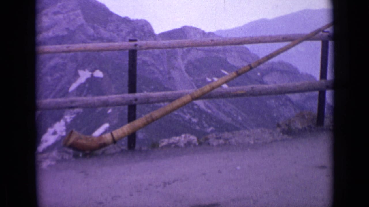 1962 Switzerland frame with a long wooden horn angled across a mountain overlook railing, with rocky peaks and snow patches beyond.