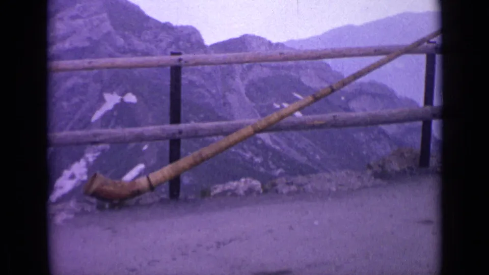 1962 Switzerland frame with a long wooden horn angled across a mountain overlook railing, with rocky peaks and snow patches beyond.