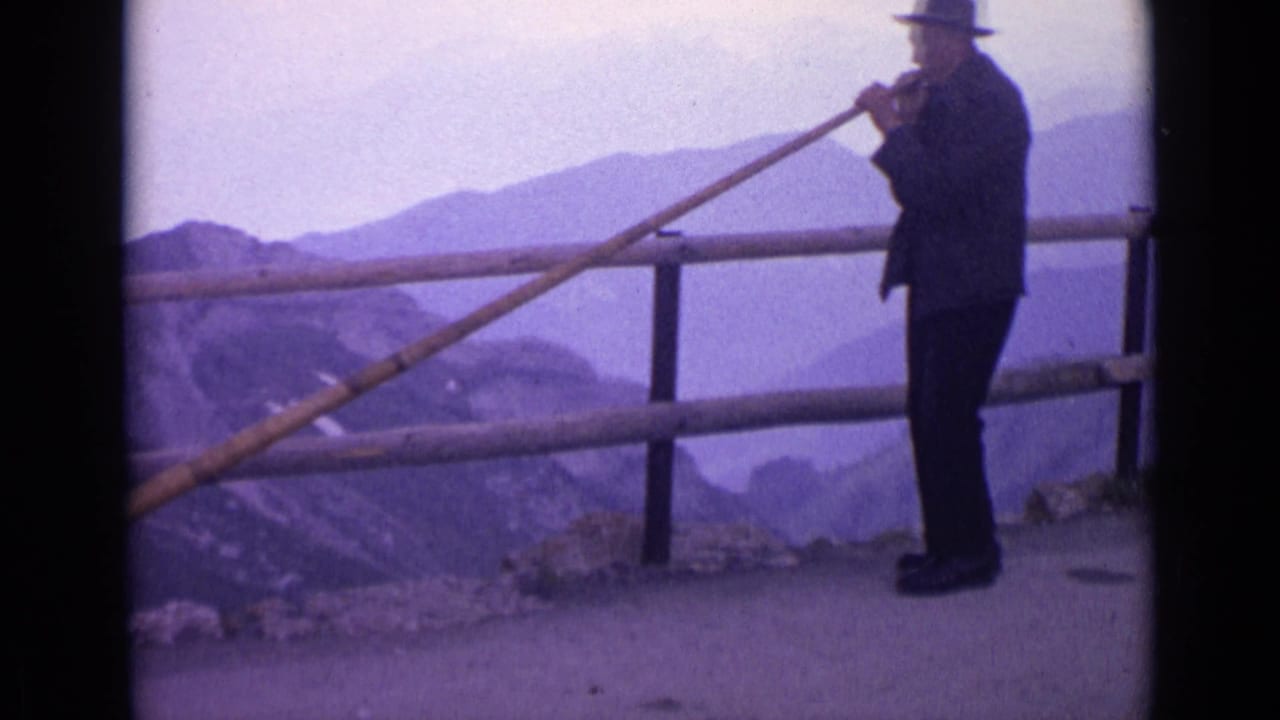 1962 Switzerland frame showing a man playing a long wooden horn at an alpine overlook, with railing and layered mountain ridges behind him.