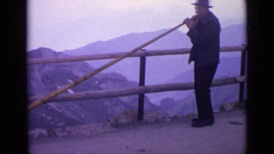 1962 Switzerland frame showing a man playing a long wooden horn at an alpine overlook, with railing and layered mountain ridges behind him.