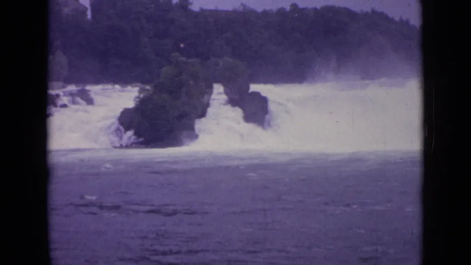 1962 Swiss 8mm frame showing white water rushing around a tree-covered rock formation with mist above the falls.