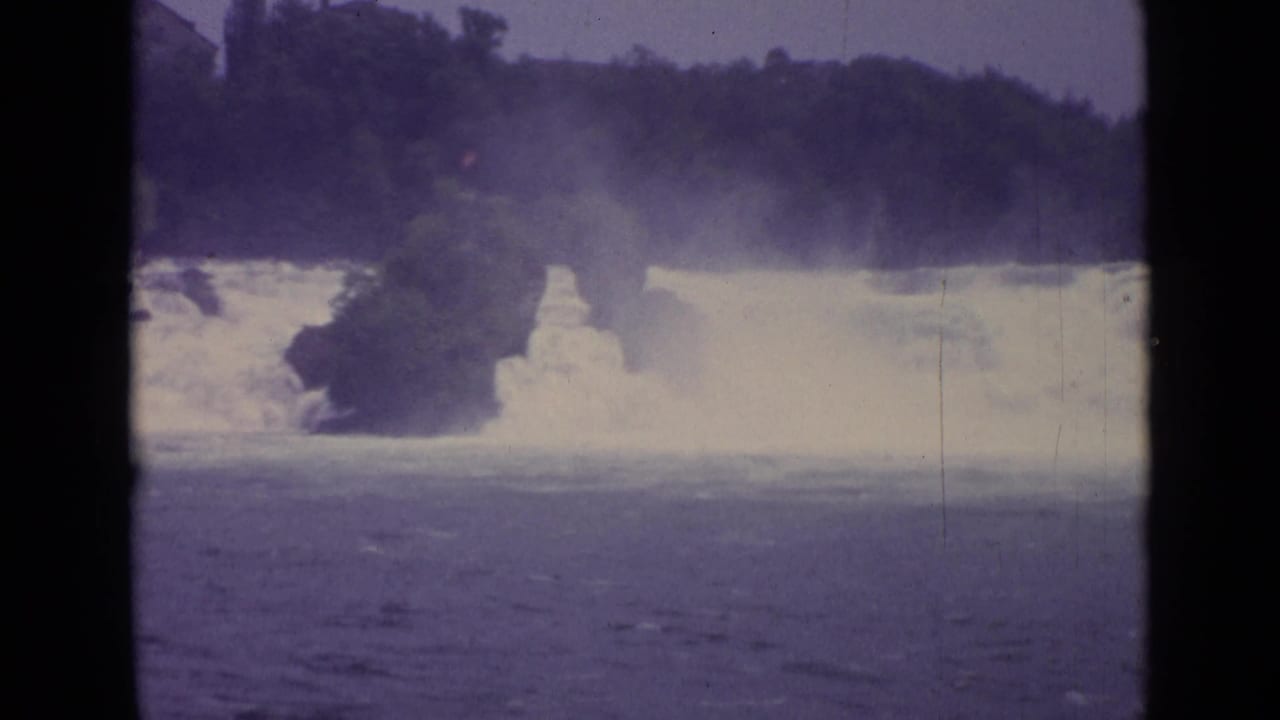 Final 1962 Swiss 8mm frame with turbulent water, mist, a rocky outcrop, and dark trees along the far bank.