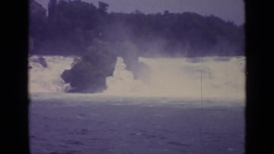 Final 1962 Swiss 8mm frame with turbulent water, mist, a rocky outcrop, and dark trees along the far bank.