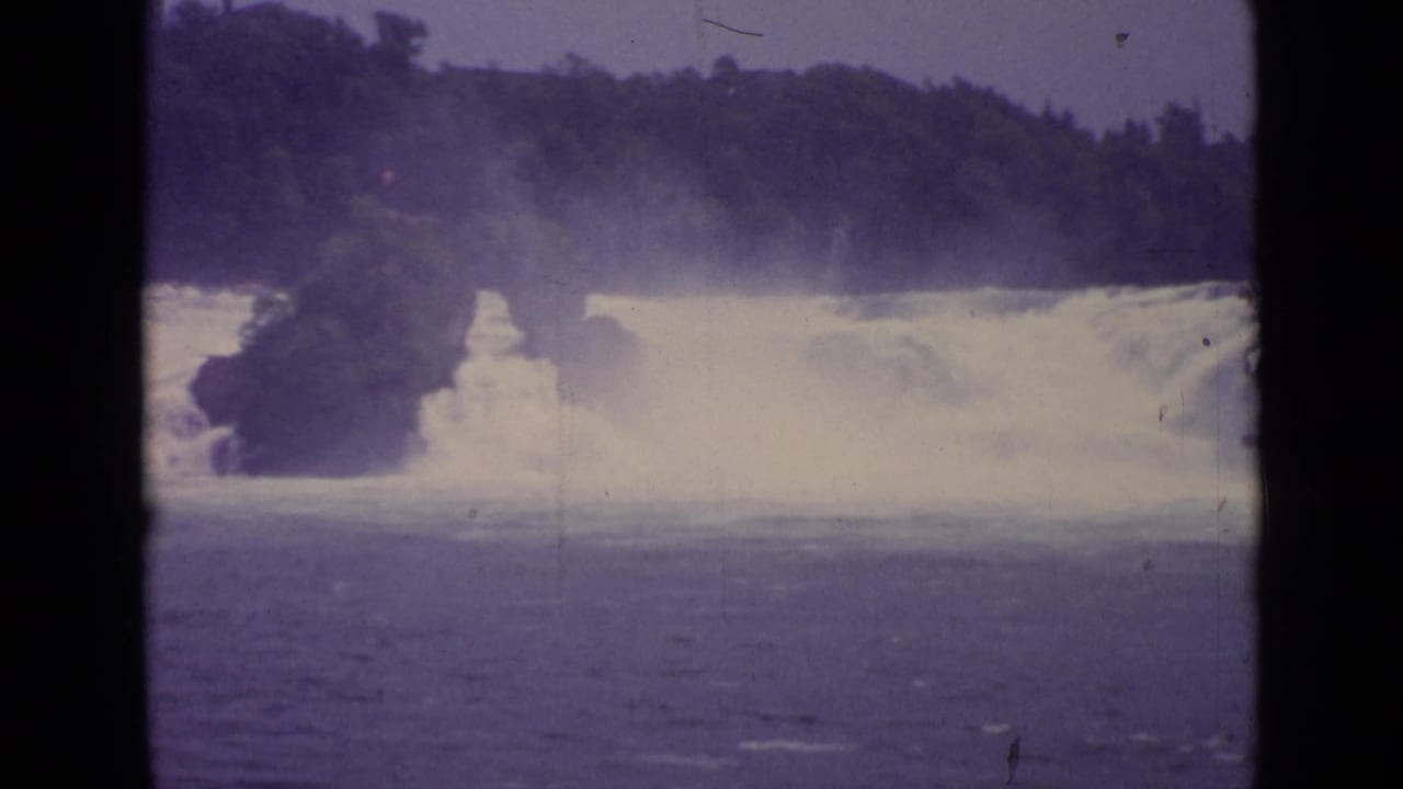 1962 Switzerland 8mm frame showing a broad waterfall pouring into river water beneath a wooded ridge.