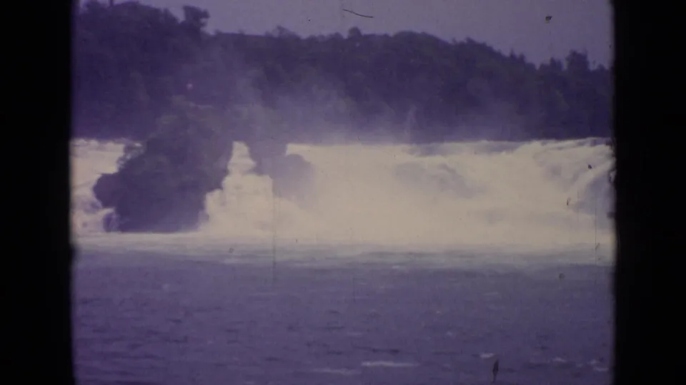 1962 Switzerland 8mm frame showing a broad waterfall pouring into river water beneath a wooded ridge.