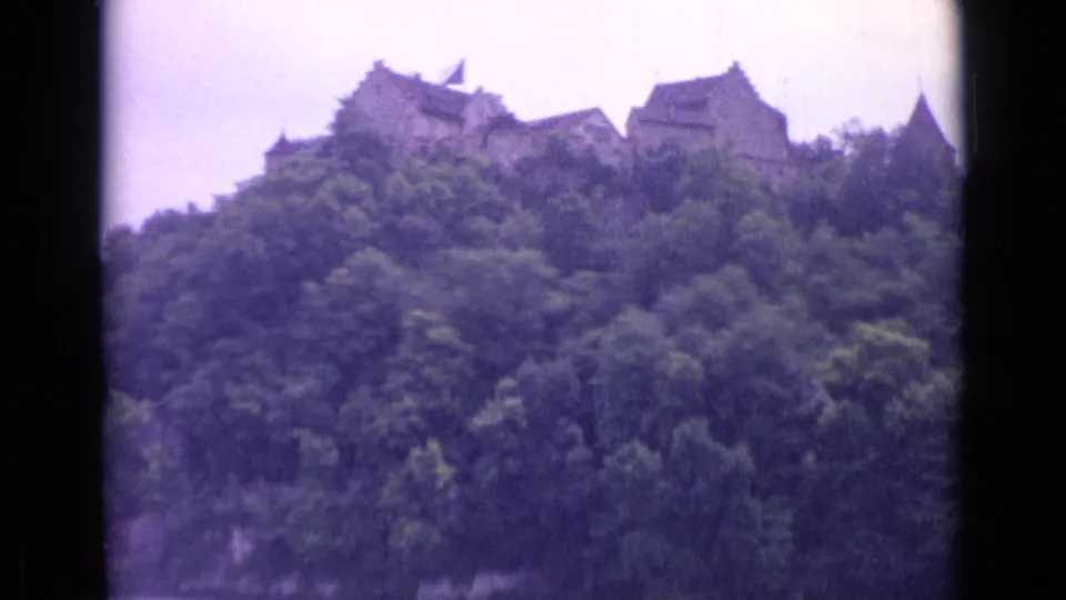 1962 Switzerland 8mm frame showing castle roofs and stone walls partly obscured by trees on a hill.