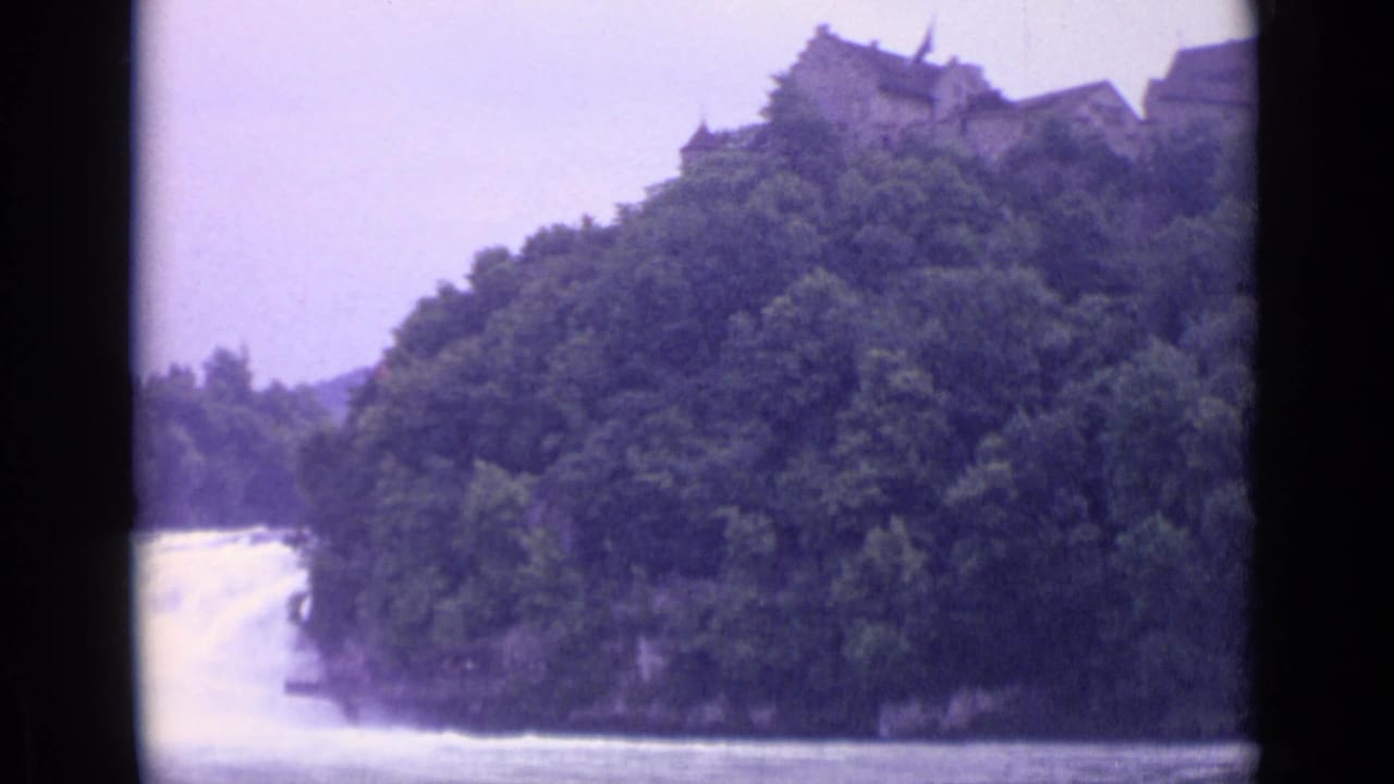 1962 Swiss 8mm frame showing a river waterfall below a wooded slope and a distant ridge-top stone structure.