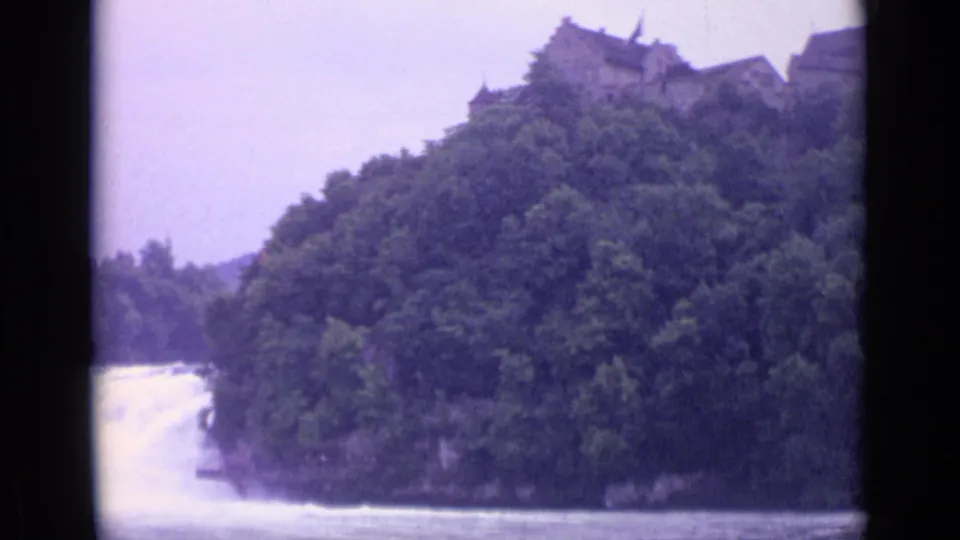 1962 Swiss 8mm frame showing a river waterfall below a wooded slope and a distant ridge-top stone structure.