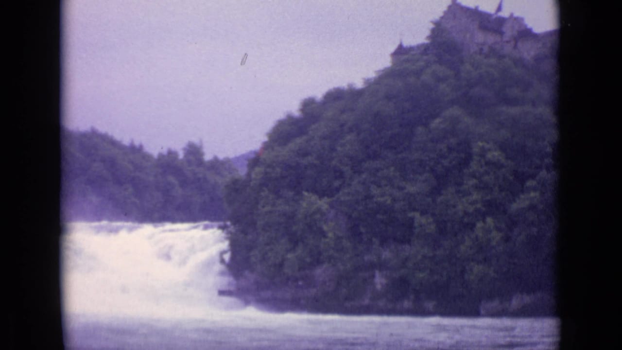 1962 Swiss 8mm view of a powerful waterfall framed by forested banks and a ridge-top stone structure.
