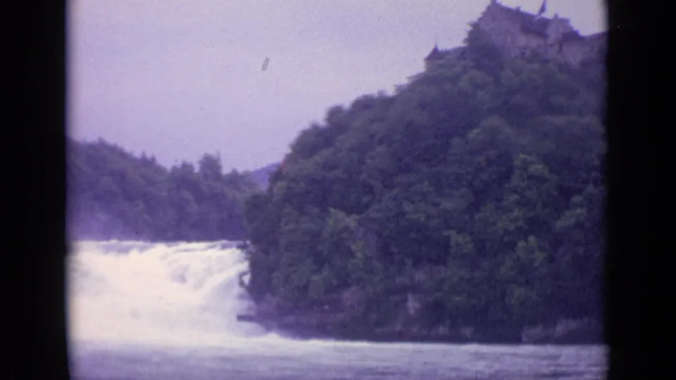 1962 Swiss 8mm view of a powerful waterfall framed by forested banks and a ridge-top stone structure.