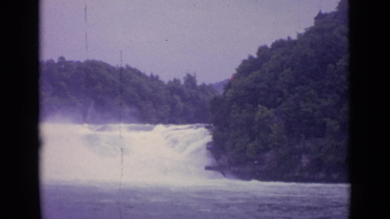 1962 Swiss 8mm frame of a river waterfall running between tree-covered banks under a pale sky.