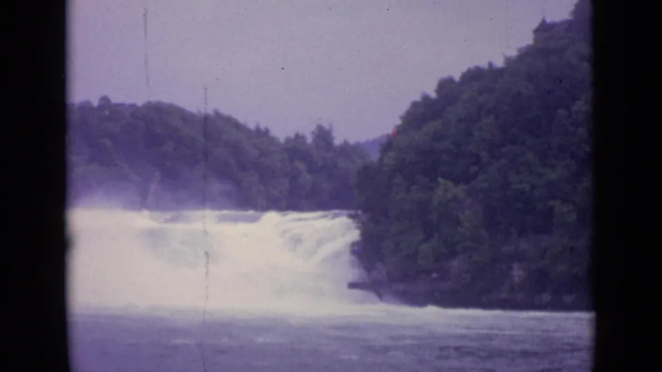 1962 Swiss 8mm frame of a river waterfall running between tree-covered banks under a pale sky.
