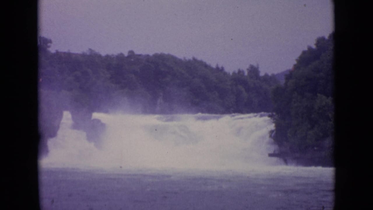 1962 Swiss 8mm frame showing a misty waterfall spanning the river between wooded hillsides.