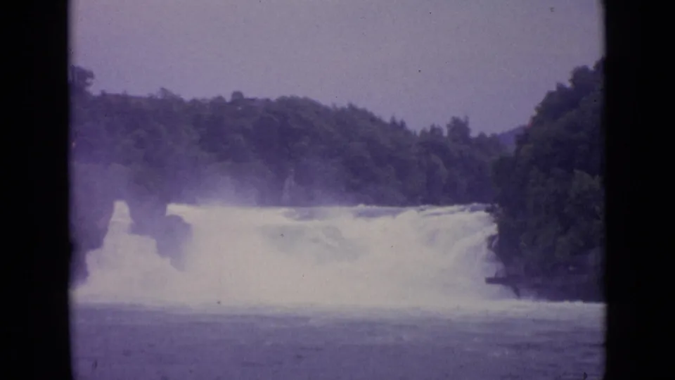 1962 Swiss 8mm frame showing a misty waterfall spanning the river between wooded hillsides.