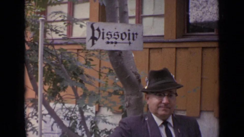 1962 Switzerland frame of a suited man in an Alpine-style hat standing outdoors below a visible Pissoir sign.