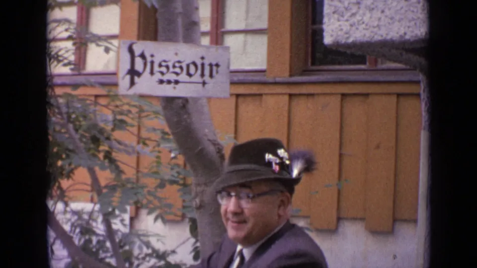 1962 Switzerland frame showing a smiling suited man in a feathered hat with a Pissoir sign visible above him.