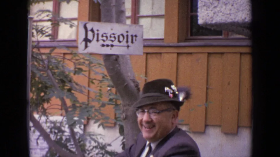 1962 Switzerland home-movie frame of a smiling man in a suit and Alpine-style hat near a white Pissoir sign.