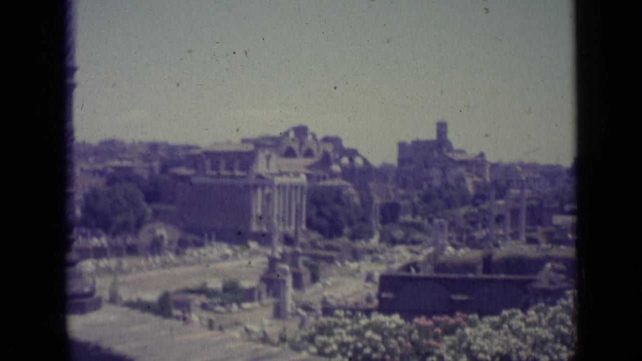 Wide view over ancient ruins with columns, open ground, and distant buildings.