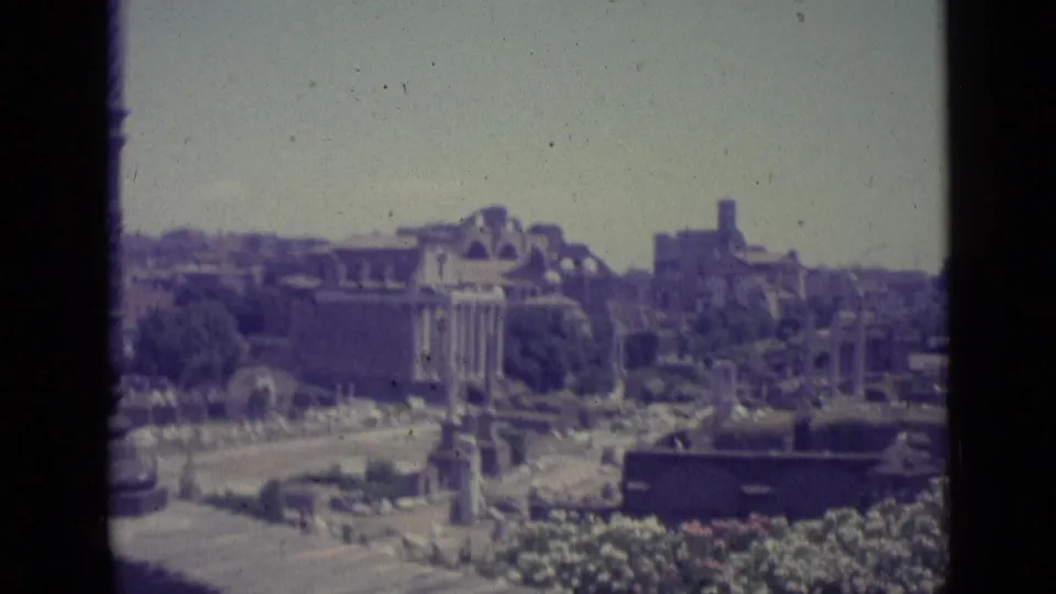 Faded 1962 8mm travel frame showing a wide Rome-area ruins view with columns, open ground, and distant hillside buildings.