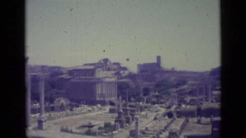 Faded 1962 8mm frame of Rome-area ruins with standing columns, paths, and distant buildings under a bright sky.
