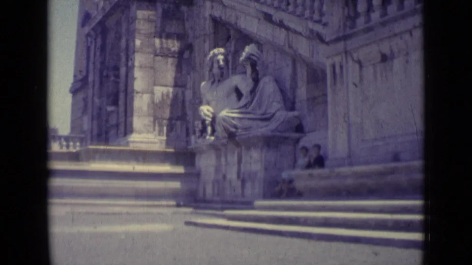 1962 Vatican City home-movie frame of a seated stone sculpture near broad steps, with church masonry and two visitors at the edge.