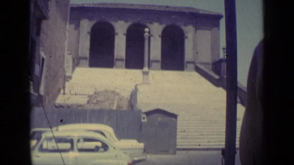 1962 Vatican City frame showing arched entries, broad stone steps, a parked mid-century car, and a pedestrian at the edge.