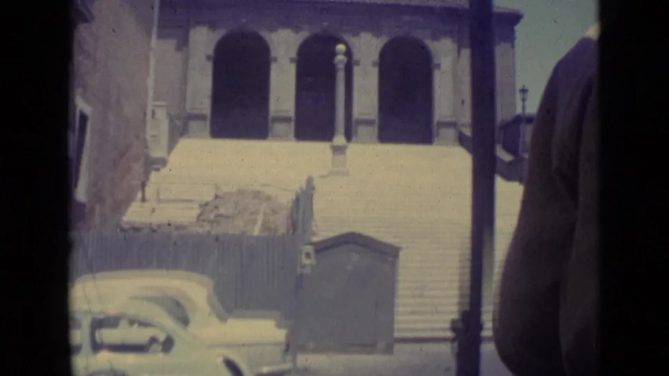 1962 Vatican City home-movie frame showing broad steps, arched facade openings, street lamps, and a parked light-colored car.