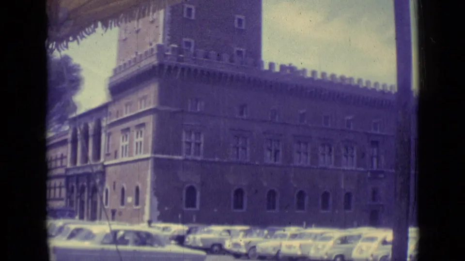 1962 Vatican City home-movie frame showing a crenellated palace facade, arched windows, and compact cars parked in front.