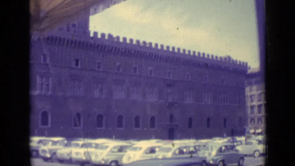 1962 Vatican City frame with a long crenellated facade, arched lower windows, and rows of parked mid-century cars.