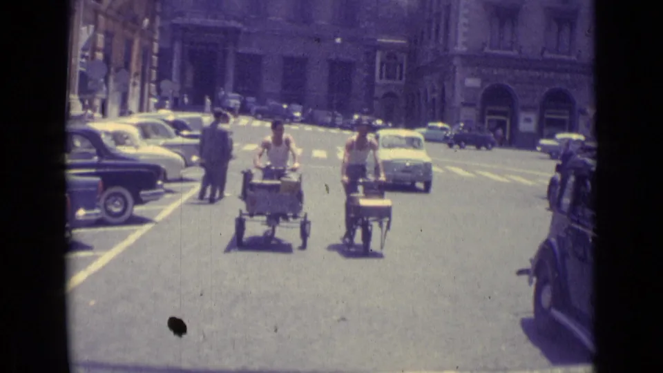 1962 Vatican City street frame showing people pushing small carts past parked and moving cars in a historic district.