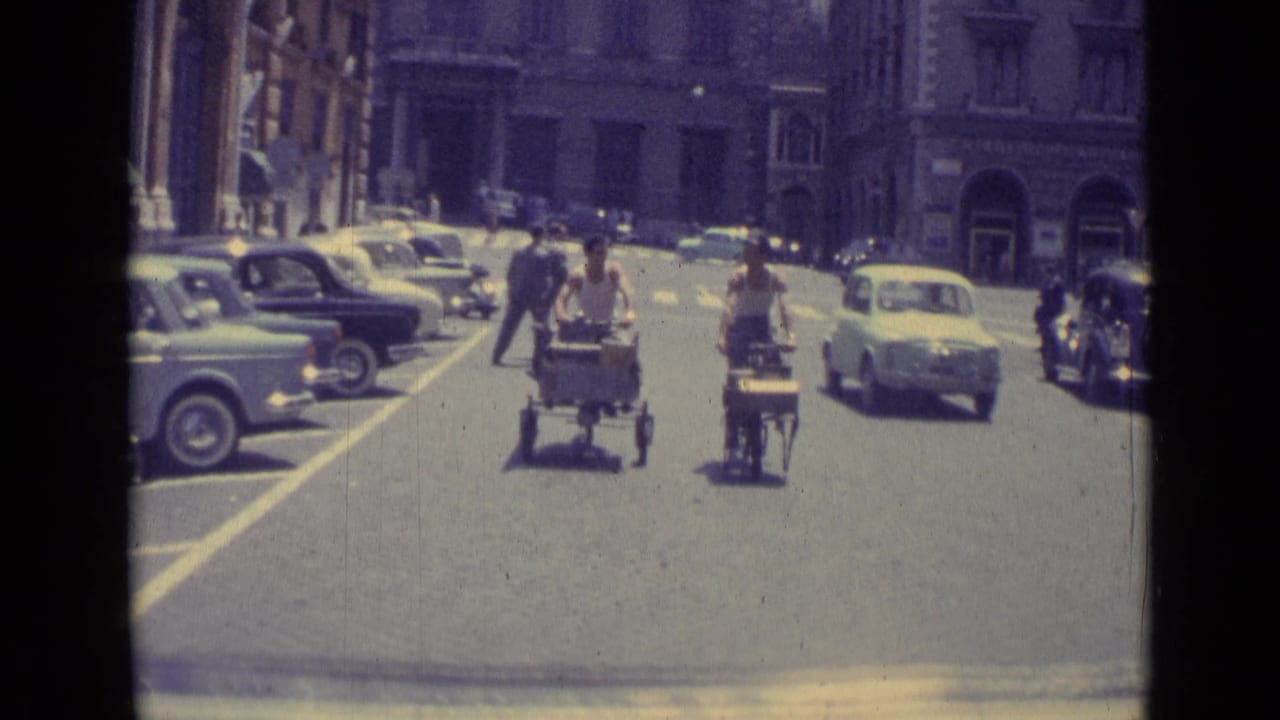 Two men ride small carts along a city street with parked and moving cars.