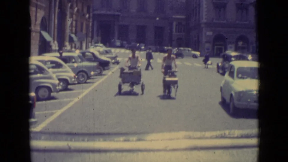 1962 Vatican City street view with two cart riders moving through traffic, compact cars lining the curb, and pedestrians crossing ahead.