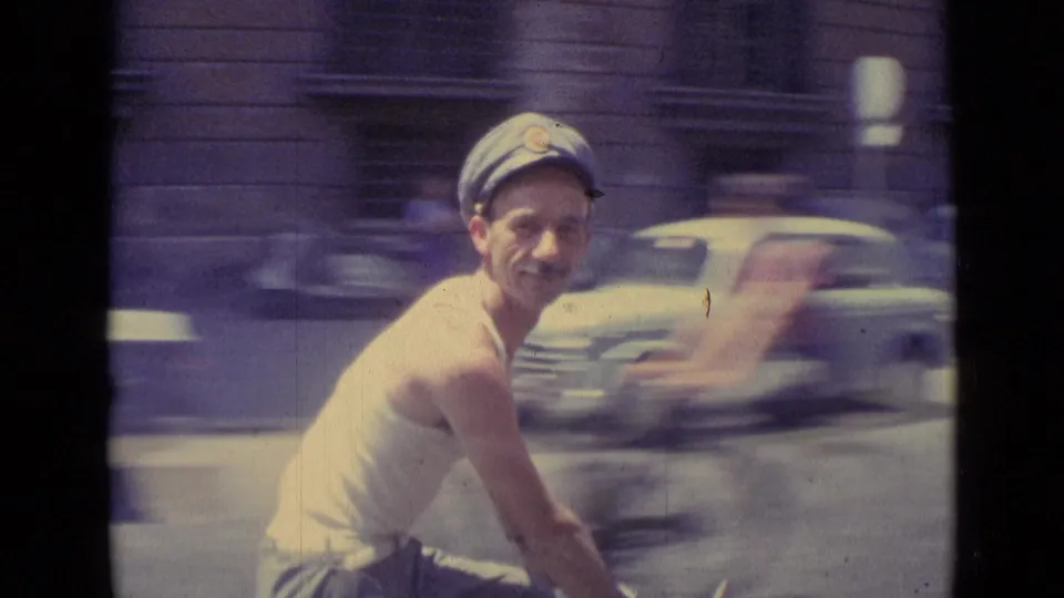Close 1962 Vatican City home-movie view of a smiling cart rider in a cap, with passing cars blurred in the background.