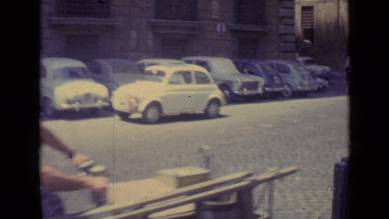 1962 Vatican City street frame with a row of compact cars, cobbled pavement, stone building fronts, and part of a rider passing near camera.