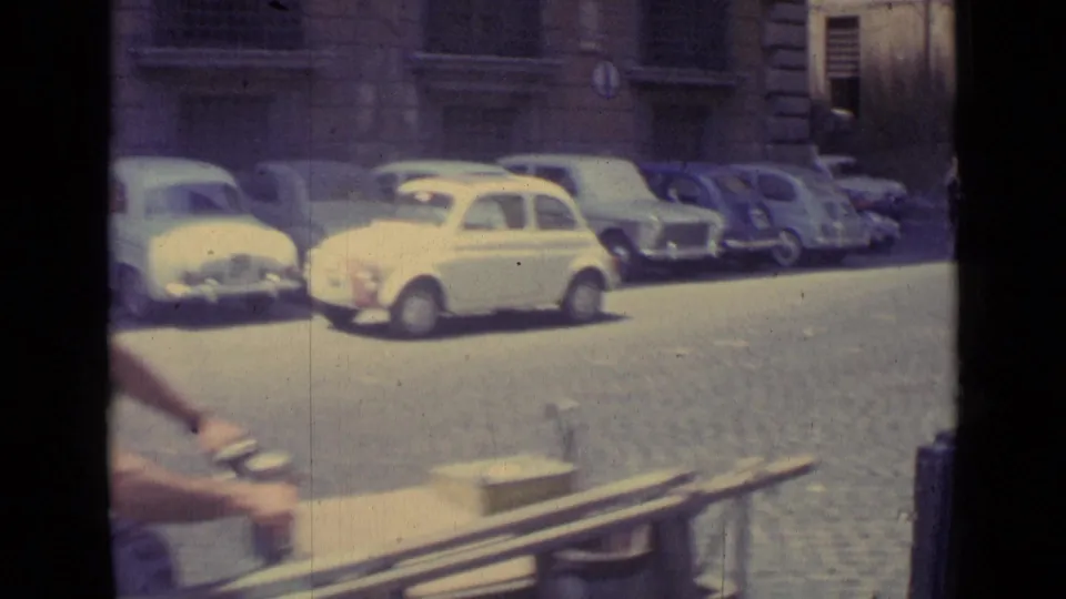 1962 Vatican City street frame with a row of compact cars, cobbled pavement, stone building fronts, and part of a rider passing near camera.