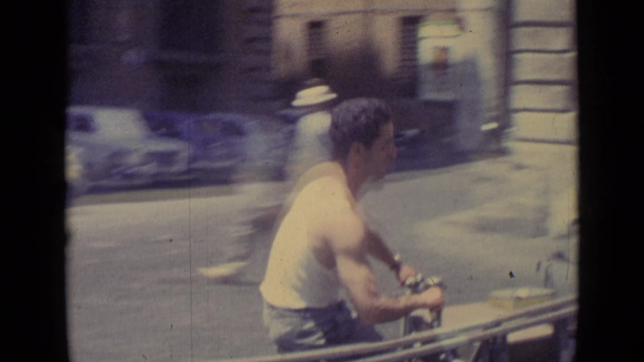 1962 Vatican City home-movie frame of a man on a motor scooter passing classic parked cars on a sunlit urban street.