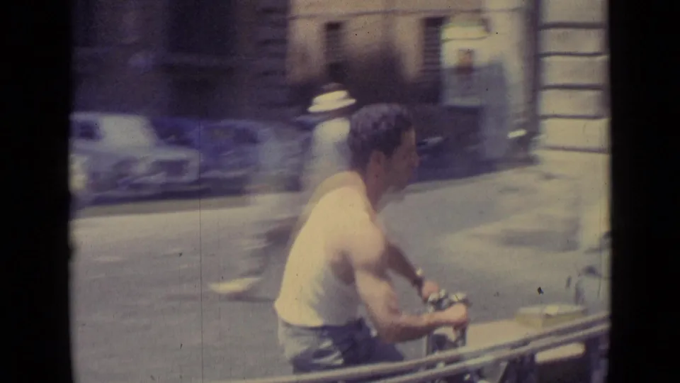 1962 Vatican City home-movie frame of a man on a motor scooter passing classic parked cars on a sunlit urban street.