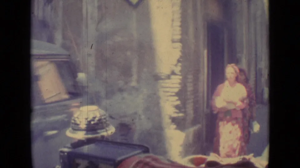 1962 Vatican City home-movie frame with a woman in a patterned dress near a sidewalk, building facade, and parked vehicle.