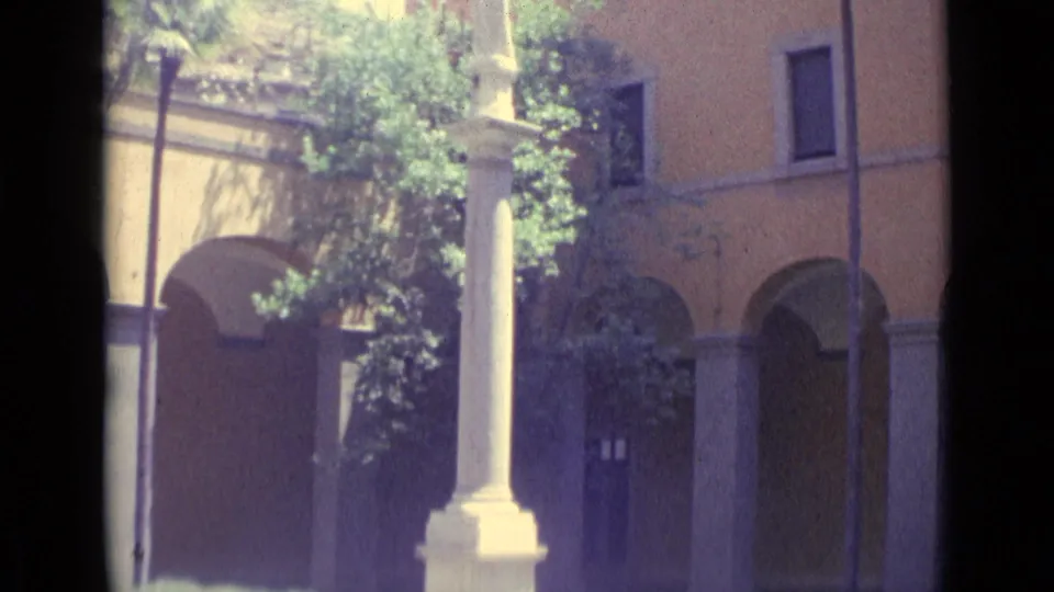 1962 Vatican City home-movie view of a tall white column before arched walls, trees, windows, and shaded courtyard planting.